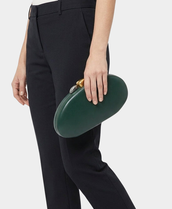 A person wearing black trousers holds a dark green oval leather clutch with a gold-tone metal clasp against a light gray studio background.